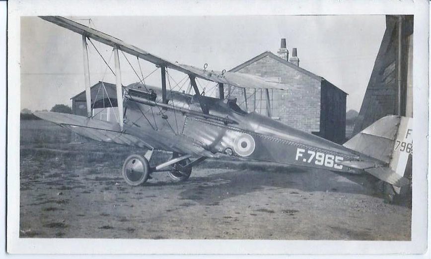 Black and whte photograph of a biplane in front of a brick building on an airfield.