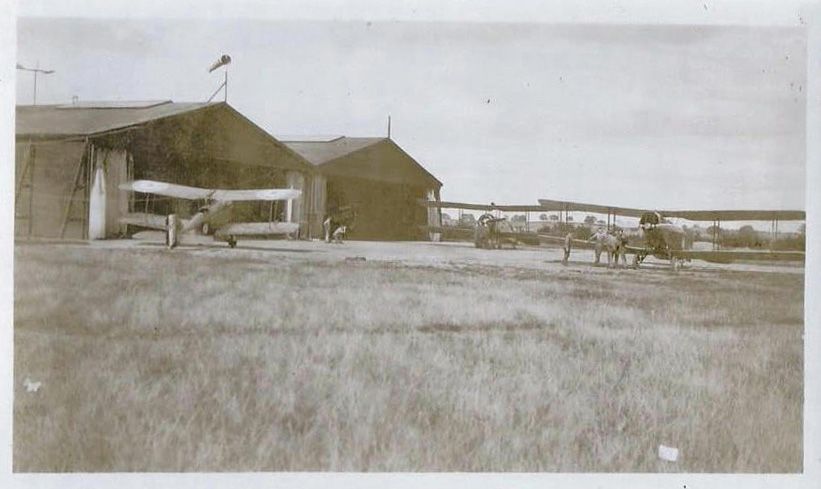 Black and whte photograph of three biplanes in front of hangars at an airfield with hills in the distance.