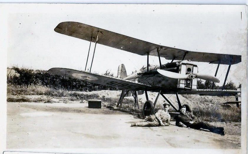 Two young men lie relaxing in front of a biplane on an airfield.