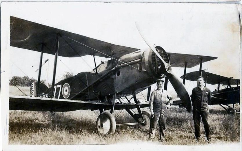 Two young men stand in front of a biplane on an airfield.