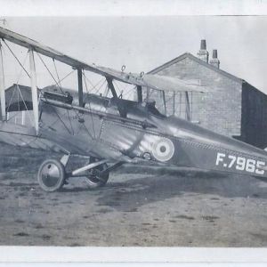 Black and whte photograph of a biplane in front of a brick building on an airfield.
