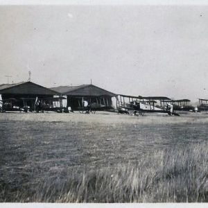 Black and whte photograph of many biplanes in front of two large hangars at an airfield.