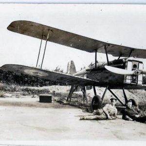 Two young men lie relaxing in front of a biplane on an airfield.